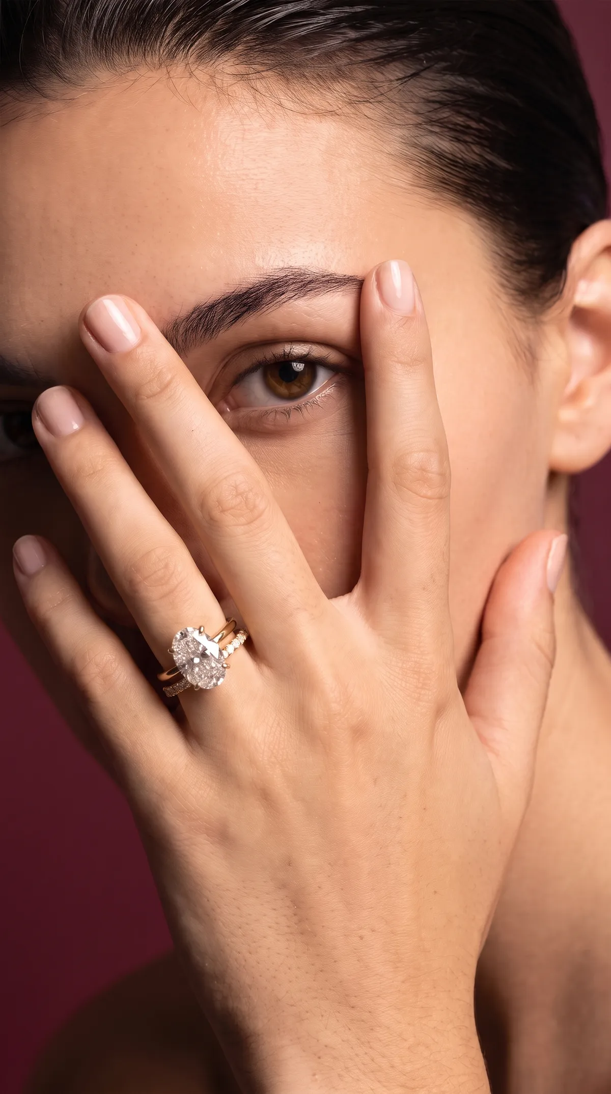 A close-up of her hand across her face, an oval diamond engagement ring stacked with a diamond eternity band