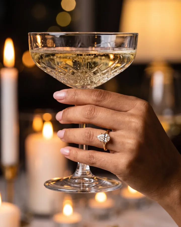 Raising a champagne coupe while wearing The Astraea pear solitaire