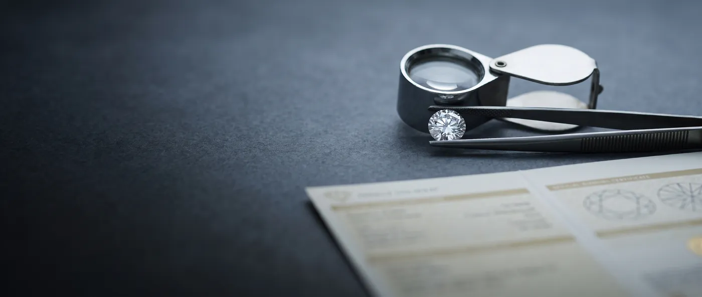 A round diamond held in tweezers beside a loupe, above a diamond grading report